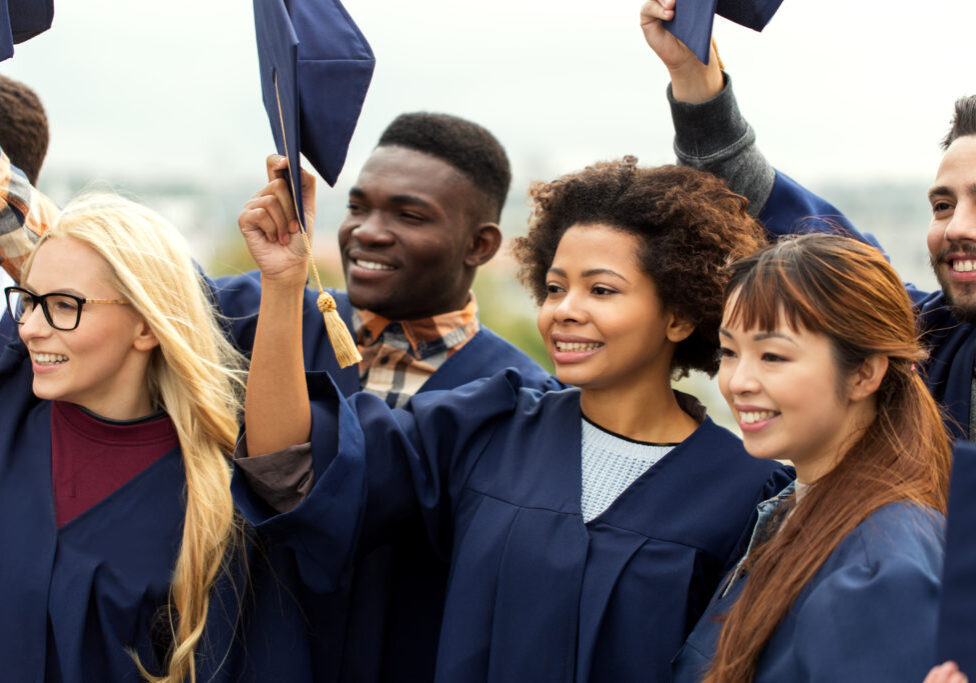 education, graduation and people concept - group of happy international students in bachelor gowns waving mortar boards